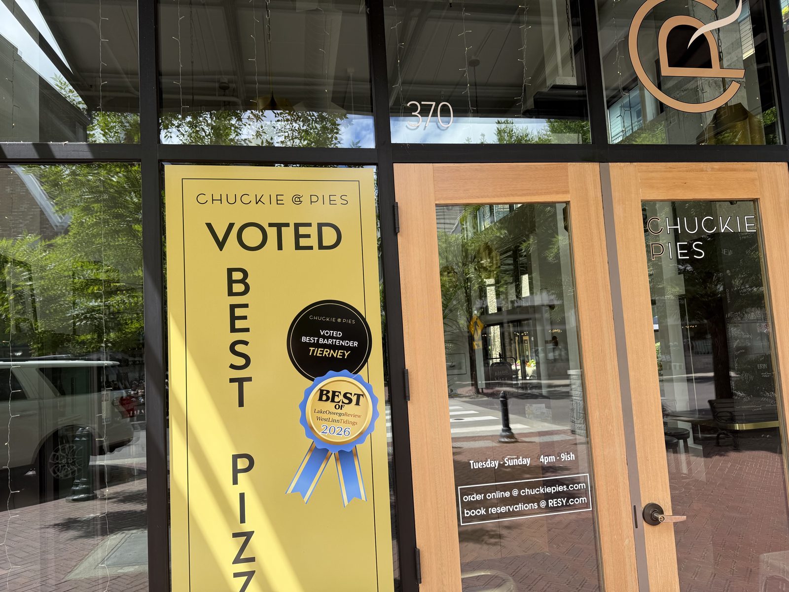 The front door of Chuckie Pies at 370 First Street in Lake Oswego, with a tall yellow poster reading Chuckie Pies, Voted Best Pizza, a black roundel for Voted Best Bartender, Tierney, and a blue and gold Best Of Lake Oswego Review and West Linn Tidings 2026 ribbon, beside a glass door lettered Tuesday to Sunday, 4pm to 9ish