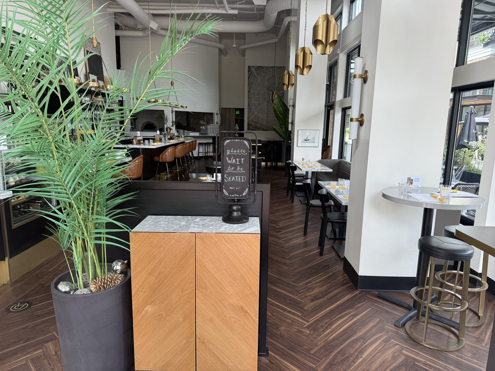 The empty dining room at Chuckie Pies photographed through the window on a Sunday midday in July, tables set with yellow napkins, a please wait to be seated chalkboard on the host stand, dark walnut herringbone floor, brass pendant lights and the wood oven visible at the back