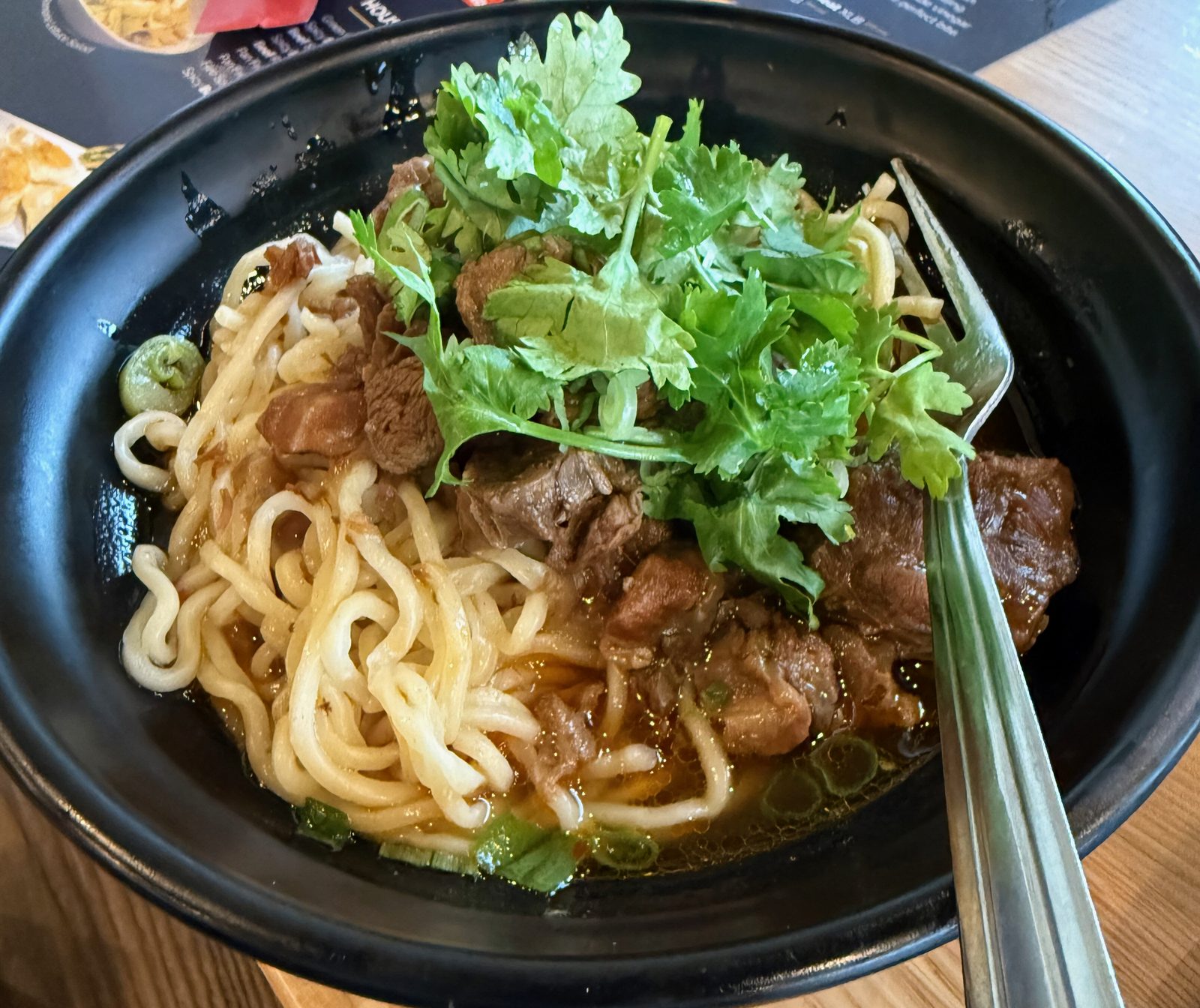 A black bowl of Dan Dan noodles at Dough Zone in Vancouver, Washington — hand-pulled noodles and braised beef under a heap of fresh cilantro, with a fork resting in the bowl