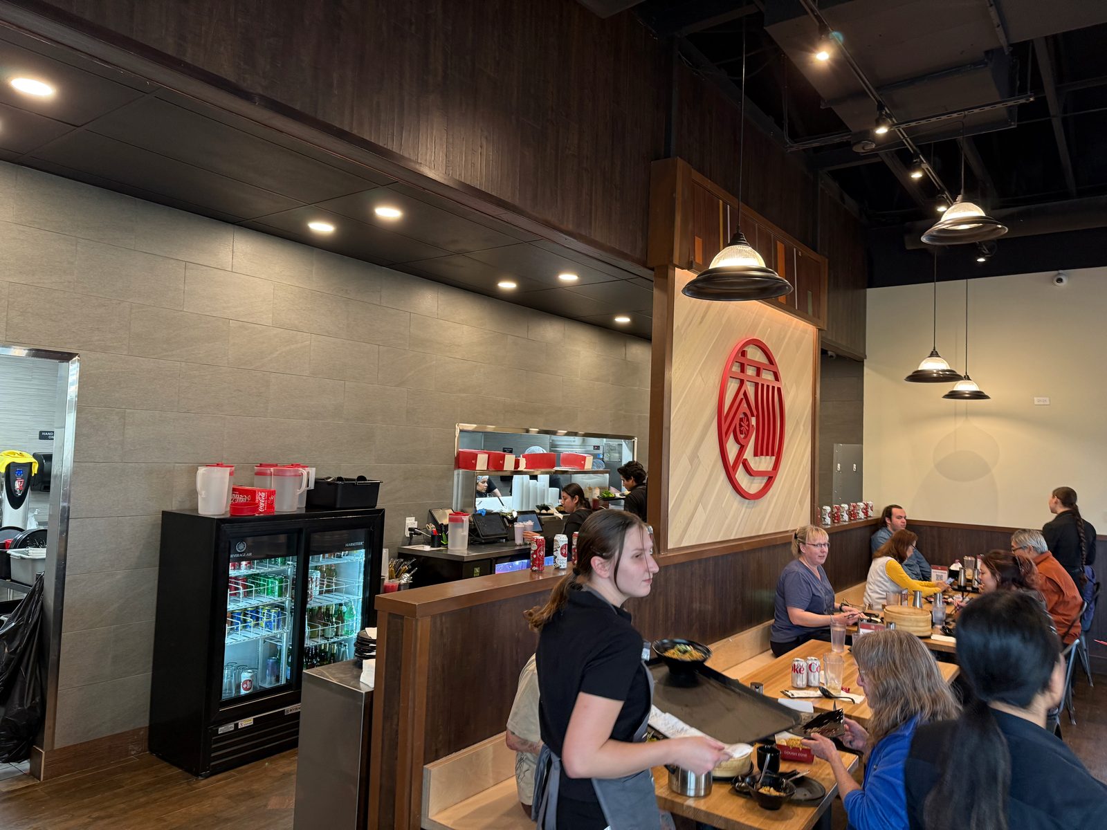 The dining room at Dough Zone in Vancouver, Washington, with servers and diners at wooden booth tables under a red circular logo medallion, and the open kitchen visible behind the counter