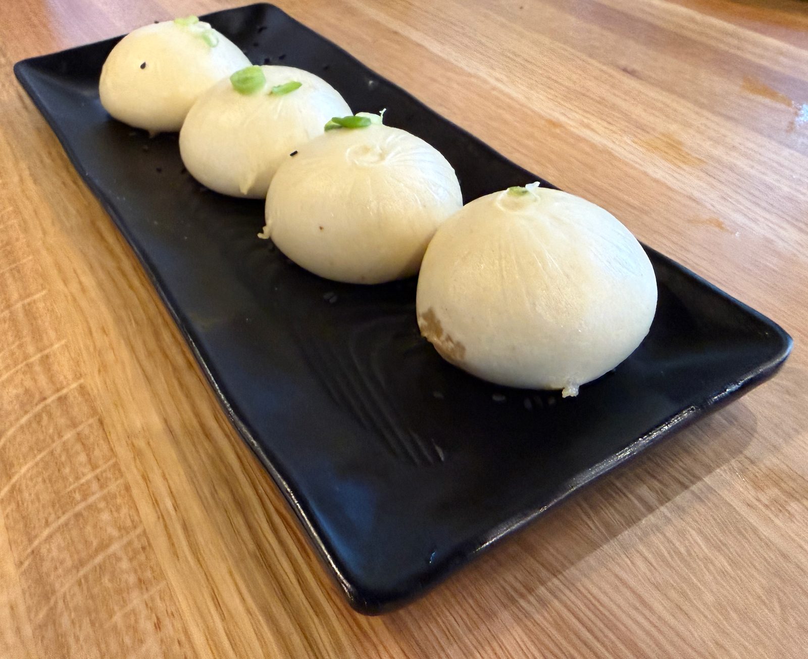 Four steamed pork buns on a black plate at Dough Zone, garnished with sliced green onion, one bitten open