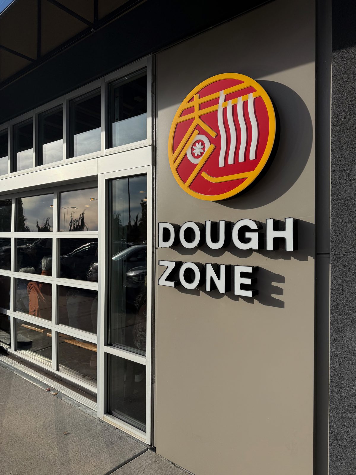 The Dough Zone sign outside its Vancouver, Washington location — a red-and-gold noodle-and-dumpling logo mark beside white DOUGH ZONE lettering on a taupe storefront panel